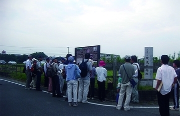 遺跡見学会を開催したときの風景写真です。参加者は説明版の前でガイドから説明を受けています。