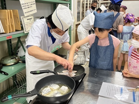 小学生が講師と一緒に魚の切り身をフライパンに入れる様子